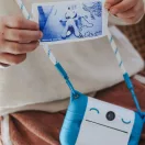 Gyermek nyakában lógó kék Kidyprint fényképezőgép a kinyomtatott képpel a kézben