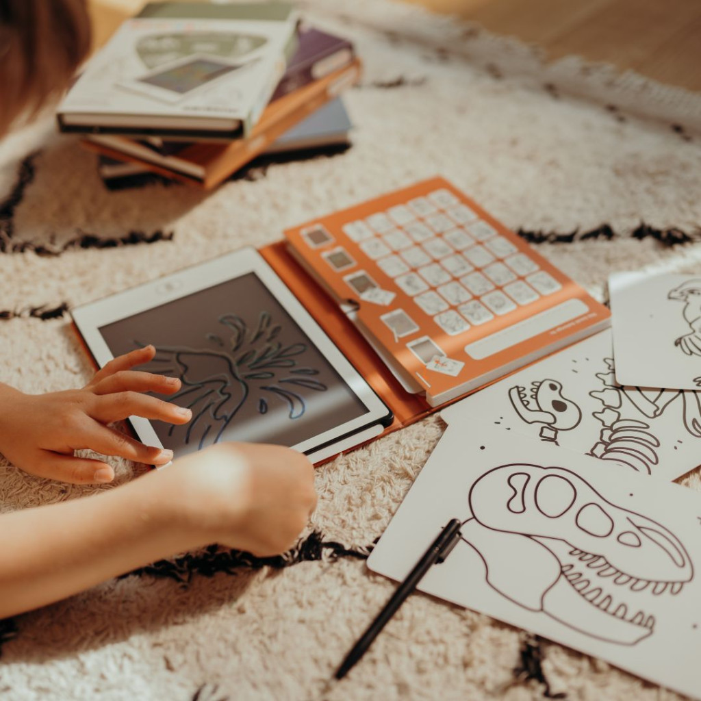 Kisgyermek rajzol a Kidywolf Kidydraw Mini LCD rajztáblára a szőnyegen, mellette a dinoszauruszos sablonkártyák